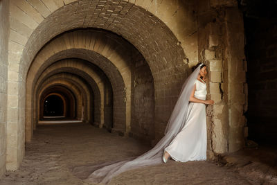 A sad elegant lady in white bride dress with a train languishes in the gloomy old castle