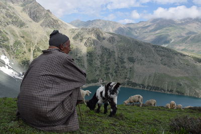 Man with sheep sitting on field against mountains