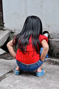 Rear view of girl crouching on footpath against wall