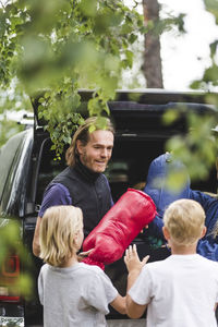 Smiling father giving luggage to children while unloading from car
