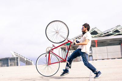Side view of man lifting bicycle on road against clear sky