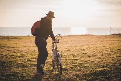 Full length of man with bicycle standing on field while looking at lake during sunny day