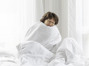 Smiling woman wrapped in blanket sitting on bed at home