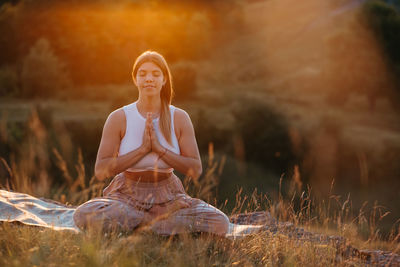 Young woman with folded hands and closed eyes sit on the mat and meditating outdoors at sunset	