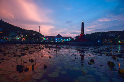 Illuminated buildings by lake against sky at sunset