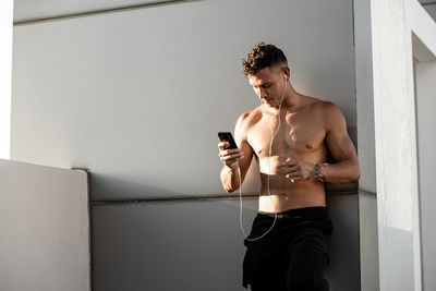 Shirtless young man listening music standing by wall