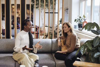 Multiracial female business colleagues discussing while sitting on sofa at coworking office