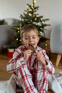 A little boy in plaid pajamas bites a gingerbread in the form of a christmas tree.