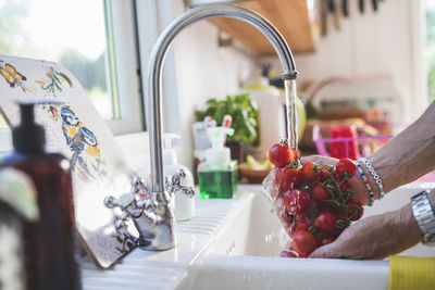 Cropped hands of man washing fresh cherry tomatoes under faucet in kitchen at home