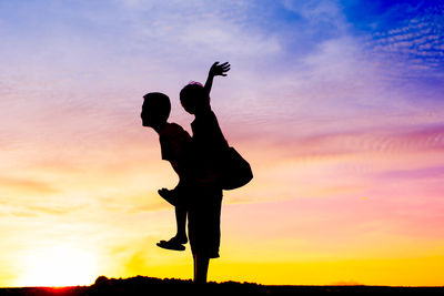 Silhouette boy piggybacking sister against orange sky