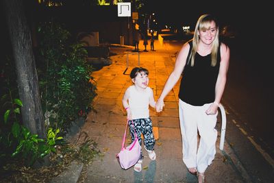 Happy woman walking with cheerful girl on sidewalk at night