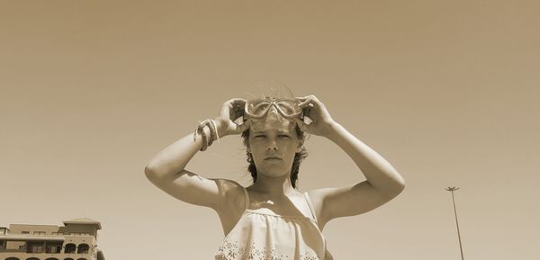 Low angle portrait of woman holding swimming goggles against sky