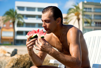 Shirtless man having watermelon against buildings