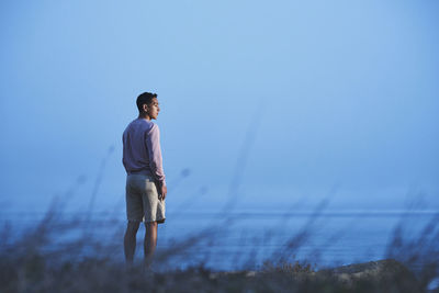 Rear view of man looking away while standing at beach against clear blue sky at dusk