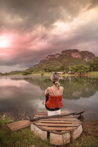 Rear view of man sitting on lake against sky during sunset