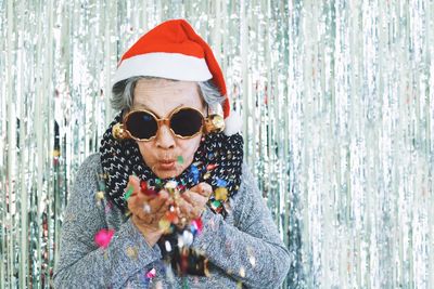 Portrait of smiling woman wearing sunglasses blowing confetti while standing against wall