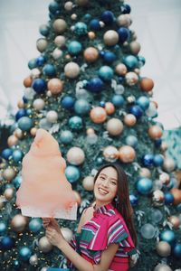 Portrait of smiling young woman standing by christmas tree holding cotton candy outdoors