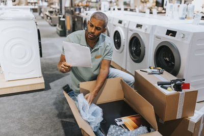 Male sales clerk reading document while opening cardboard box in appliances store