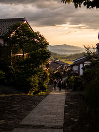 People walking on road amidst buildings against sky during sunset