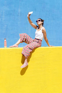 Trendy woman sitting on yellow wall while taking a selfie