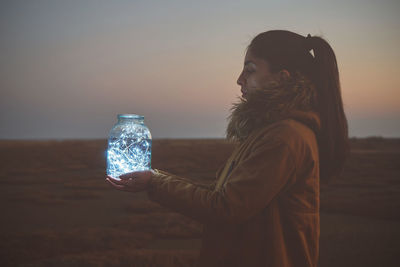 Side view of woman holding lights in jar against sky during sunset