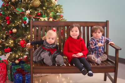 Cute babies and girl sitting on bench against christmas tree at home