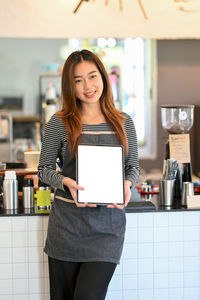 Young asian female barista holding, showing blank tablet screen mockup 