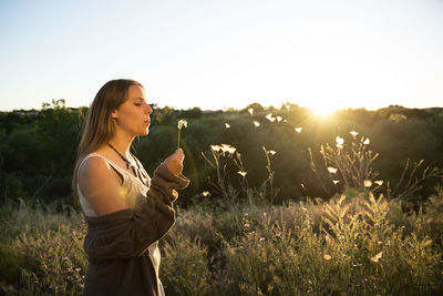 Woman blowing dandelion while standing against sky during sunset