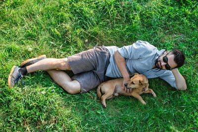 Attractive european man in sunglasses is lying on grass and hugging his cute little dog.