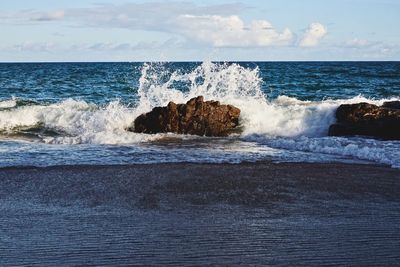 Waves splashing on rocks at shore against sky