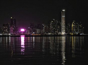 Illuminated buildings by river against sky at night