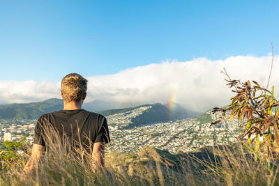 Rear view of man looking at landscape while sitting on mountain against sky