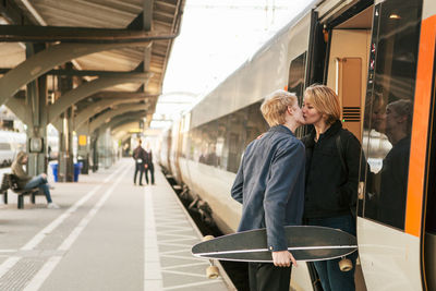 Young man holding skateboard while kissing teenage girl at doorway of train