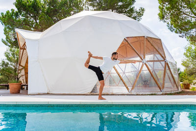 Side view of anonymous barefoot male standing in natarajasana pose near swimming pool and tent while practicing yoga