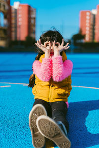 Girl forming binoculars with fingers while sitting on sports court during sunny day