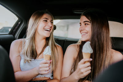 Cheerful millennial female friends eating ice cream cone in car during road trip in summer day