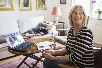 Happy senior woman looking away at home while dog relaxing on sofa