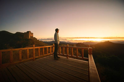 Man standing on railing against sky during sunset