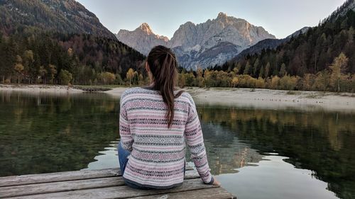 Rear view of woman sitting on pier by lake against mountains