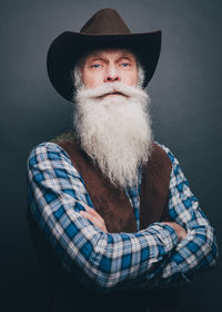 Bearded senior man wearing cowboy hat standing arms crossed against gray background