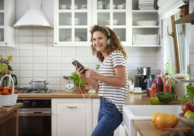 Side view of cheerful teenage girl listening music in kitchen at home