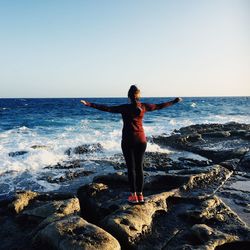 Rear view of woman standing on rock at beach against clear sky