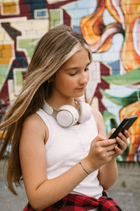 Teen girl with headphones posing on colorful city graffiti, using smartphone, looking at screen