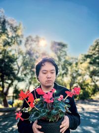 Portrait of young asian man holding red flowering cyclamen potted plant against trees and blue sky