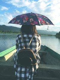 Rear view of woman holding umbrella while sitting in boat on lake against sky