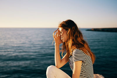 Young woman sitting by sea against sky during sunset