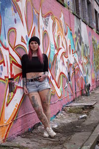 Portrait of tattooed woman with pink hair leaning on graffiti wall