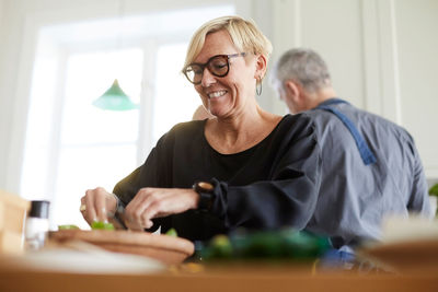 Smiling mature woman preparing food with friend in background at home