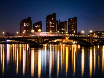 Battersea bridge and river reflections with silhouetted buildings in the background at night.