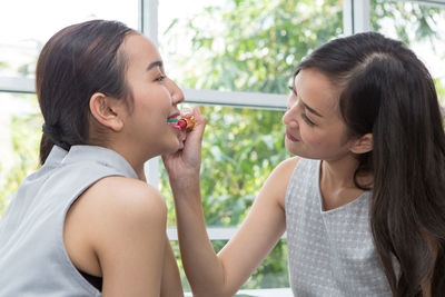 Young woman applying lipstick to sister by window at home